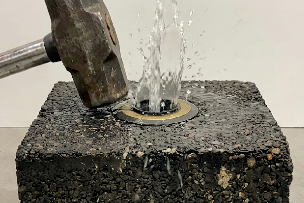 A hammer striking an asphalt block to simulate traffic loading, causing trapped water to immediately eject vertically out of the PaveVent™ relief insert.